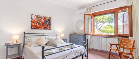 A bedroom with a bed, bedside tables, a chair, a dresser, a window with curtains, and a wall-mounted air conditioning unit.
