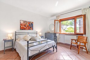 A bedroom with a bed, bedside tables, a chair, a dresser, a window with curtains, and a wall-mounted air conditioning unit. - Villa Adriana Capalbio With A/c, BBQ and Parking (Capalbio)