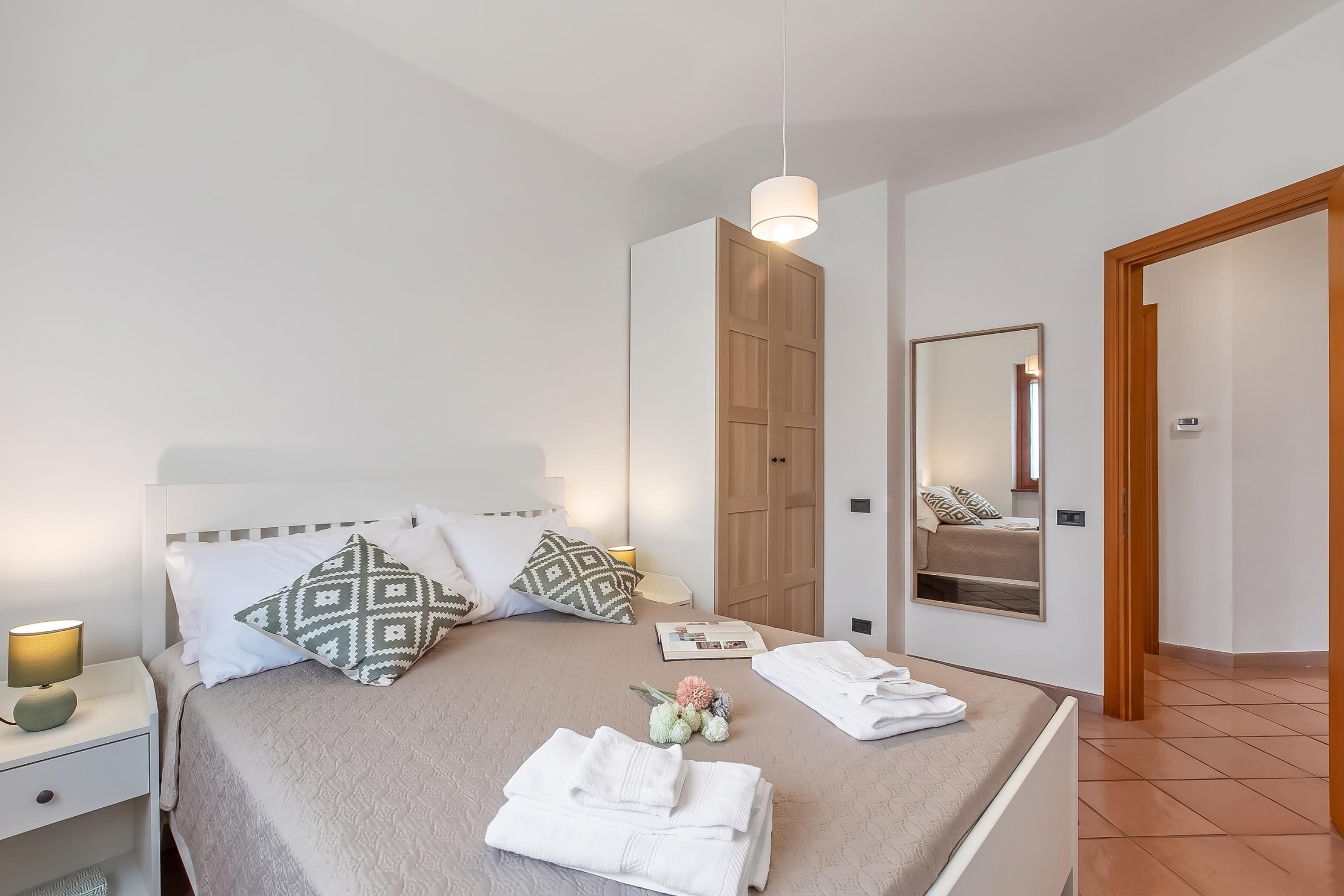 Een net opgemaakt bed met kussens, een bedlampje, een spiegel en een deur naar de badkamer.