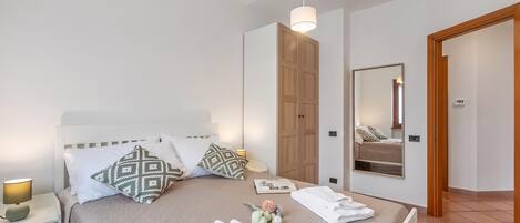 Een net opgemaakt bed met kussens, een bedlampje, een spiegel en een deur naar de badkamer.