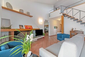 A modern living room with a sofa, a flat-screen TV, a fireplace, a staircase, and a kitchen in the background. - Villa Adriana Capalbio (Capalbio)