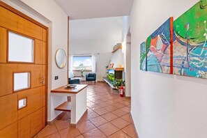 Un couloir avec une porte en bois, une petite table sur laquelle se trouve un plateau, une chaise bleue, un canapé, un téléviseur et un grand tableau abstrait.