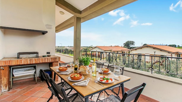 An outdoor dining area with a grill, table set with plates, glasses, and bottles, overlooking a residential area. - Villa Adriana Capalbio With A/c, BBQ and Parking (Capalbio)