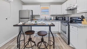 Eine moderne Küche mit weißen Schränken, einem Edelstahl-Kühlschrank, einer Mikrowelle, einem Backofen und einem Gasherd. An einem zentralen Tisch stehen zwei Hocker mit Geschirr und Tassen.