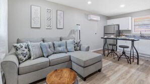 A modern living room with a grey sectional sofa, a wooden coffee table, and a kitchen area with white cabinets and a dining table.