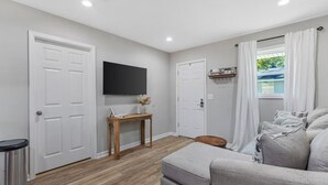 A room with a grey sofa, a wooden table with a vase, a TV on the wall, and a window with curtains.