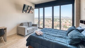 A bedroom with a large bed, a grey sofa, a desk, and a city view through large windows. - Casa Grace a pasos de Polanco y Reforma. (Ciudad de México)