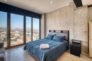 Ein Schlafzimmer mit einem großen Bett, einem Nachttisch, Blick auf eine Stadtlandschaft durch große Fenster und einer Betonwand.