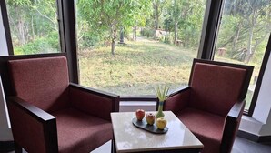 Une pièce avec deux fauteuils rouges, une petite table sur laquelle se trouvent un vase et des pommes, et une vue sur un jardin par de grandes fenêtres.