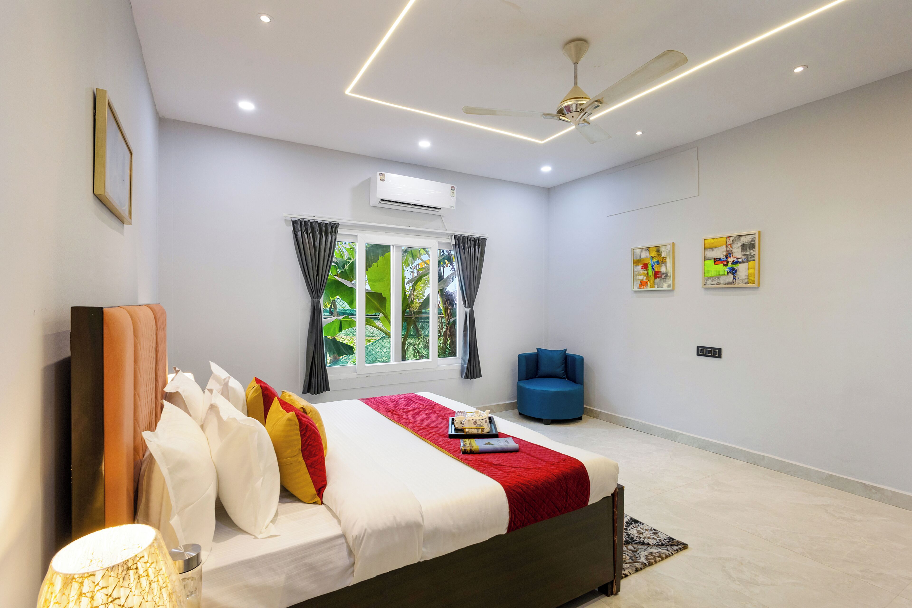 A bedroom with a bed, a ceiling fan, an air conditioner, a window with curtains, a blue chair, and artwork on the wall.