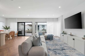 A modern living room with a sofa, a flat-screen TV, and a dining area with chairs and a table. - Beachside on Beecroft (Huskisson)
