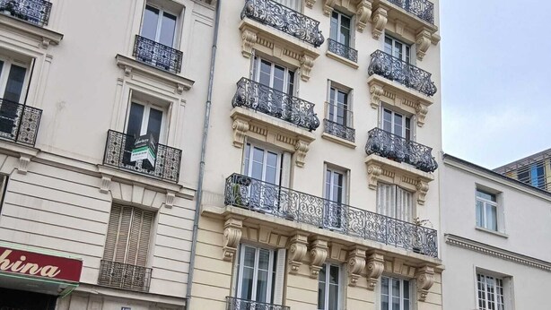 Un edifici de diversos pisos amb balcons, una botiga amb un cartell i cotxes aparcats.