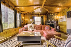 Interior acolhedor de uma cabana, com decoração em madeira, lareira de pedra e uma grande janela.