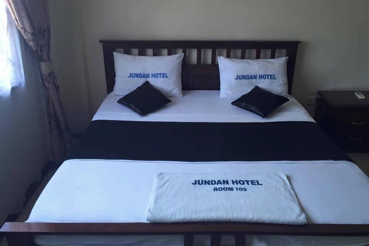 Ett hotellrum med en säng, två kuddar med texten ”JUNDAN HOTEL”, ett sängbord och en filt med texten ”JUNDAN HOTEL ROOM 109”.