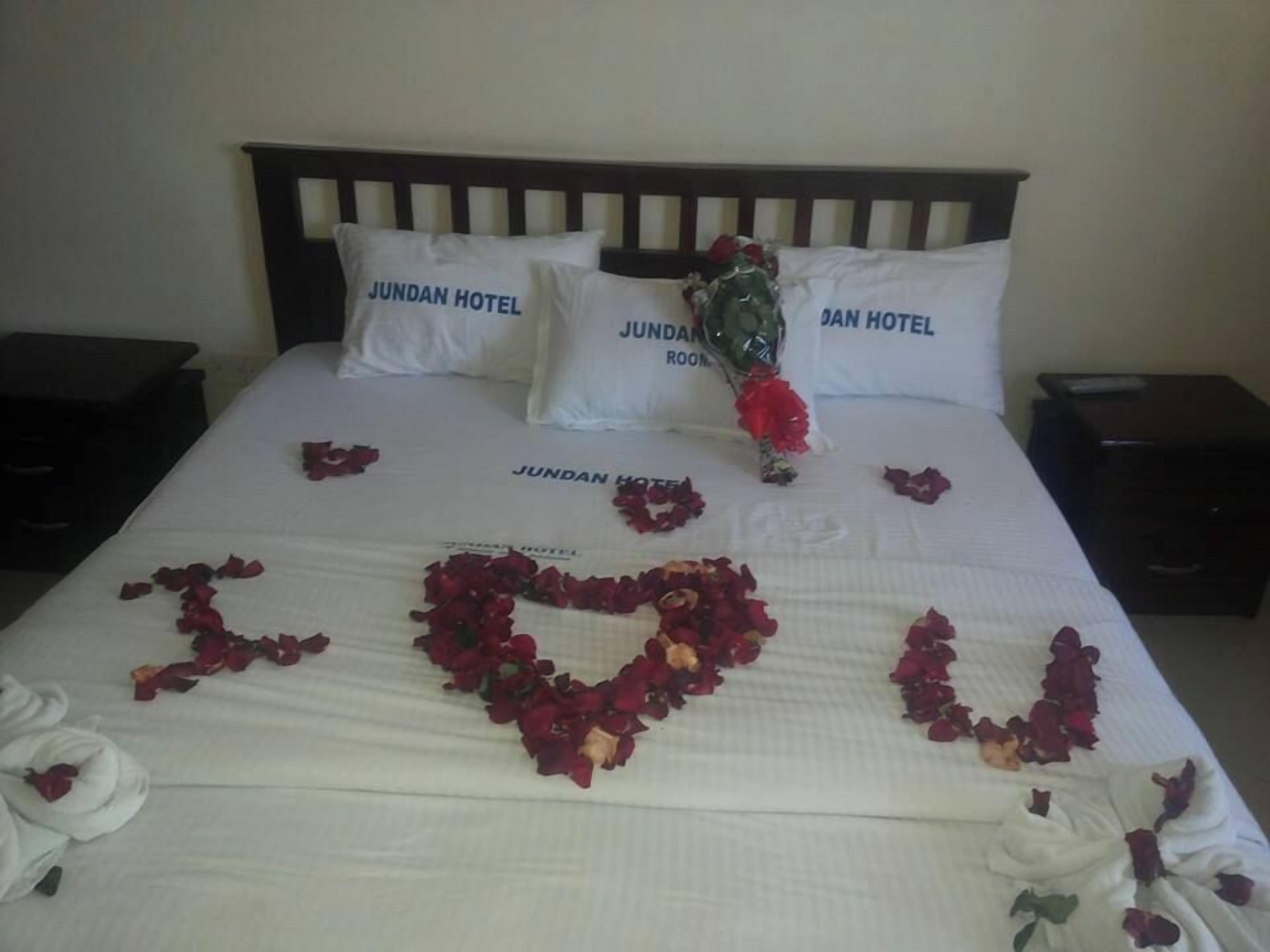 A bed with a heart made of rose petals spelling 'LOVE' and pillows labeled 'JUNDAN HOTEL ROOM'.
