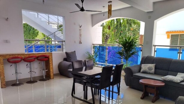 A spacious living area with a dining table, chairs, a sofa, and a small coffee table. There are three red bar stools and a large window offering a view of greenery and a blue structure outside. - Jundan Palace Hotel Mombasa (Mombasa)