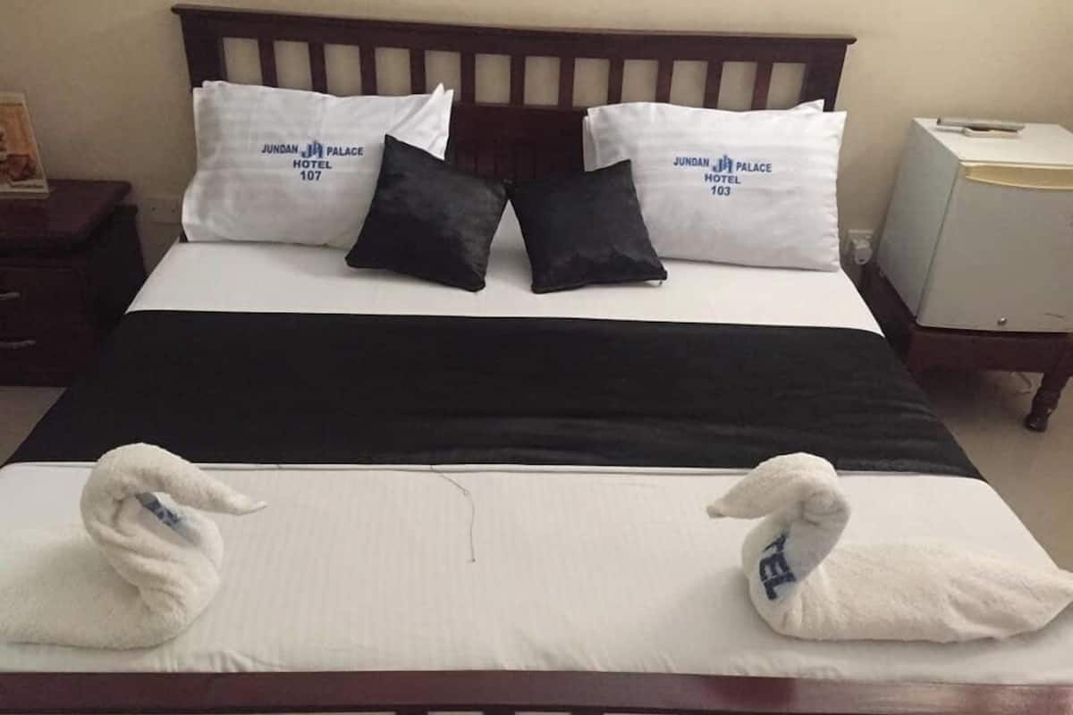 En säng med vita sängkläder, två kuddar med texten ”JUNDAN JA PALACE HOTEL”, ett svart överkast och två handdukar med symbolerna ”J” och ”J”.