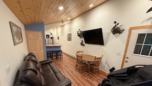 A living room with a leather sofa, a dining table with chairs, a TV mounted on the wall, and a guitar case on the floor.