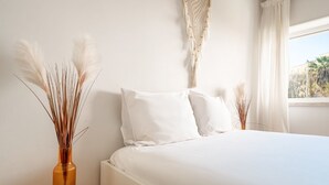 A bedroom with a bed, white pillows, a nightstand with a vase, and a macrame wall hanging. - GuestReady - Retreat in Almada (Almada)