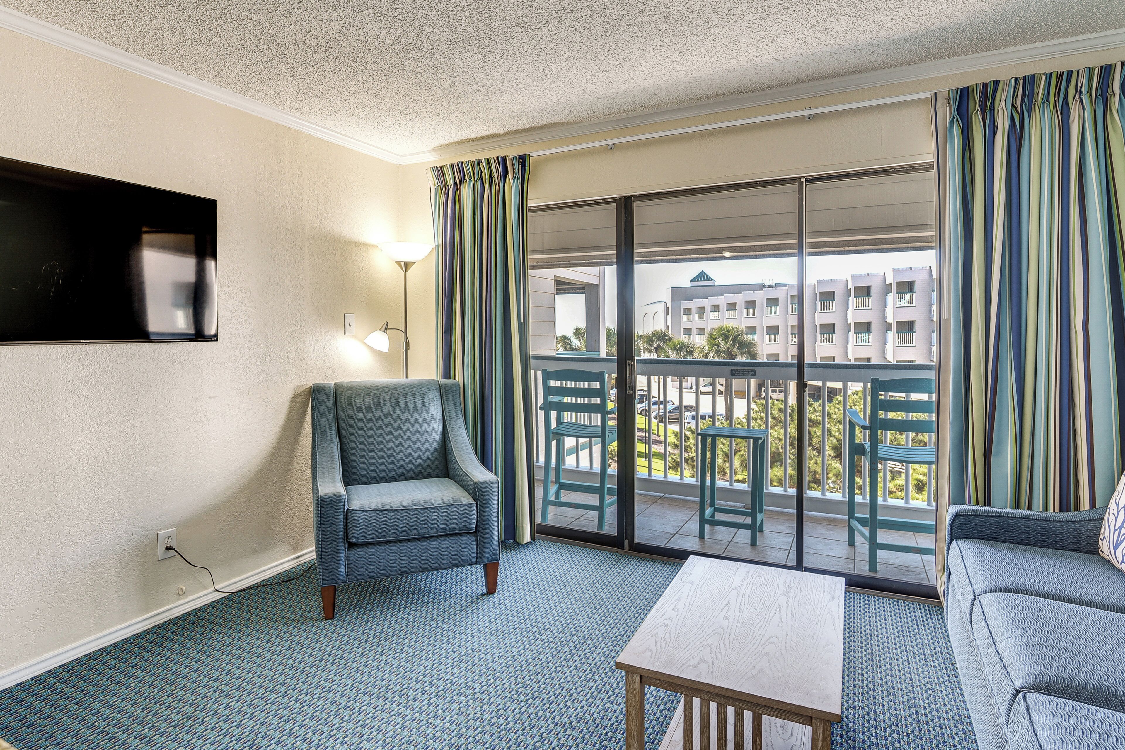 Une chambre d’hôtel avec un canapé, un fauteuil, une table, un téléviseur fixé au mur et un balcon donnant sur des immeubles.