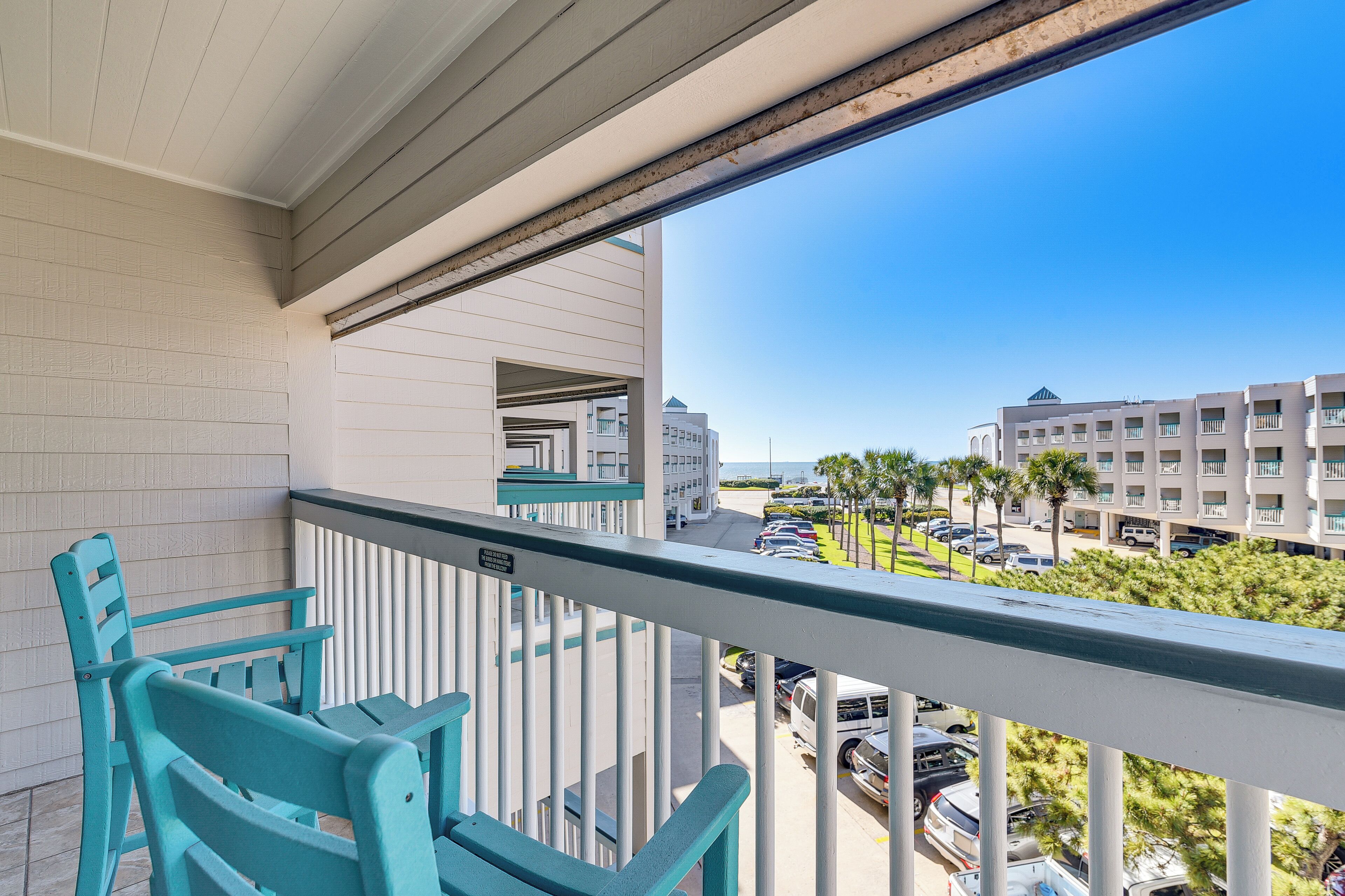 A balcony with teal chairs, a view of a parking lot, and buildings in the background.