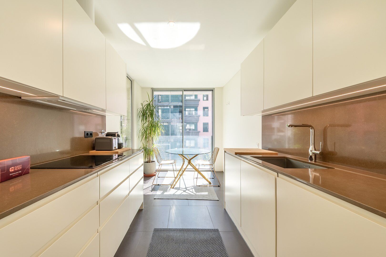 Une cuisine moderne dotée d’un îlot central, d’un coin repas avec une table en verre et d’une vue sur un immeuble grâce à de grandes fenêtres.