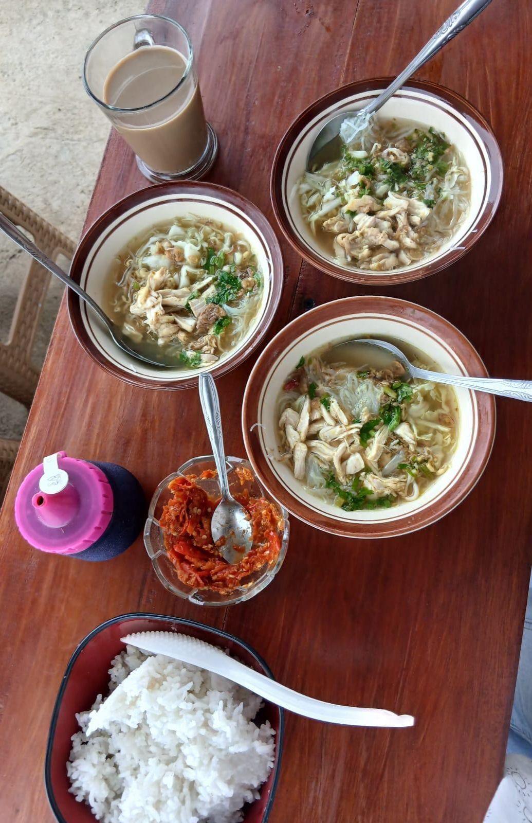 Una mesa con cuatro tazones de sopa, un vaso de té helado, un plato de arroz y un recipiente rosa con una cuchara.