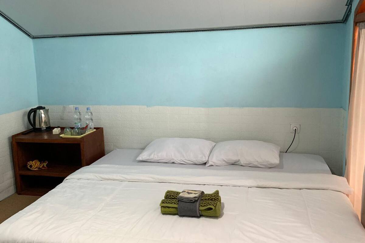 A single bed with white bedding, a wooden nightstand with a kettle and water bottles, and a power outlet on the wall.