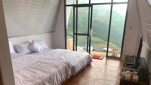 A bedroom with a large bed, wooden flooring, a desk with a computer, and a view of a green landscape through a large window.