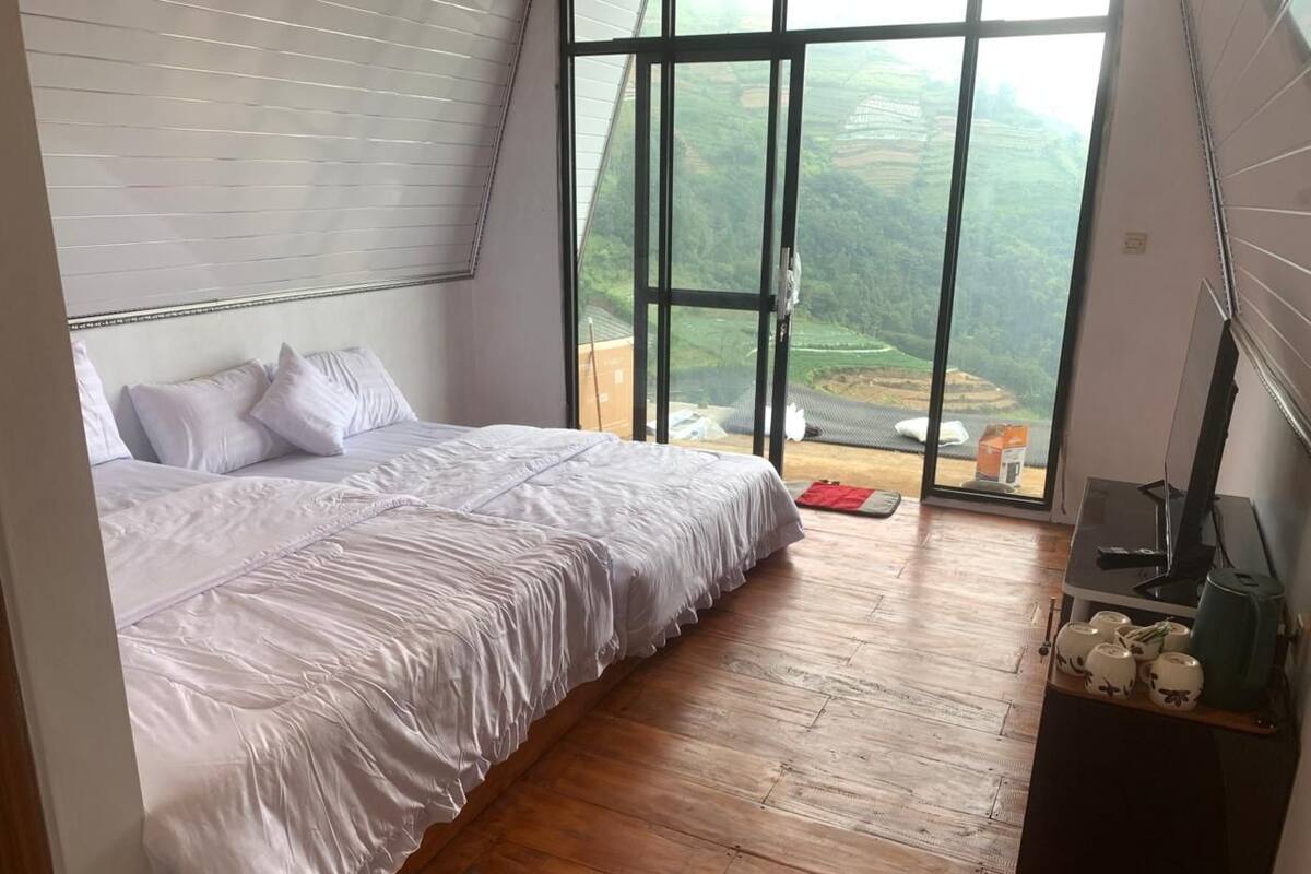 A bedroom with a large bed, wooden flooring, a desk with a computer, and a view of a green landscape through a large window.