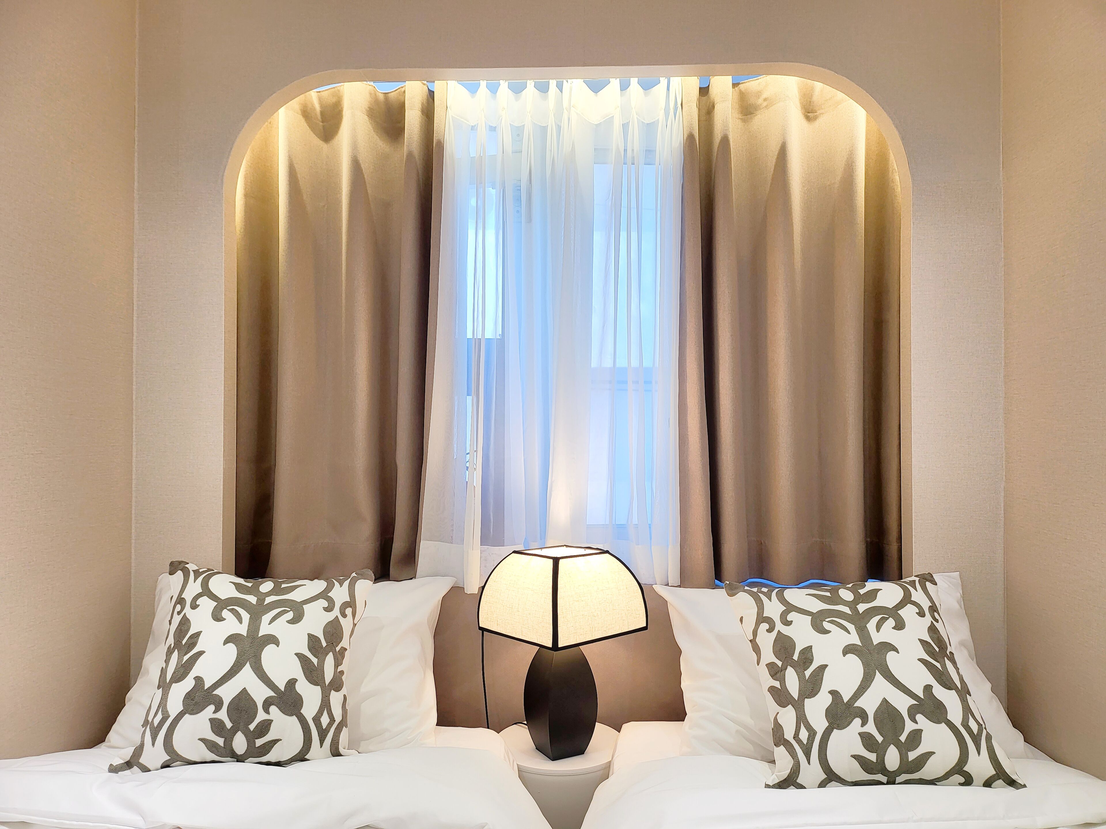Ein Hotelzimmer mit einem Bett, zwei gemusterten Kissen, einem Nachttisch mit Lampe und einem Fenster mit Vorhängen.