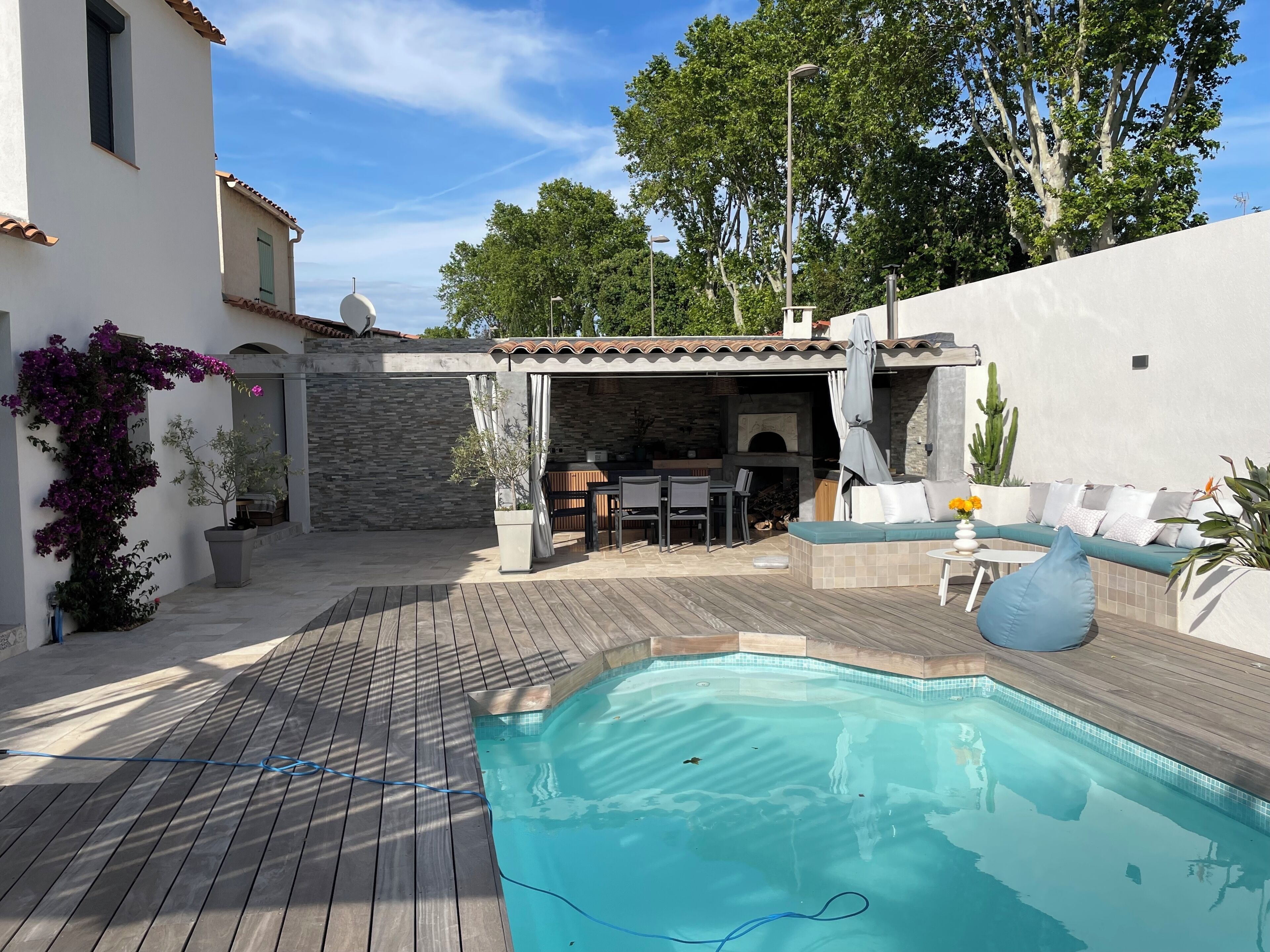 A pool with a wooden deck, a stone wall, and a covered outdoor seating area with a table and chairs.