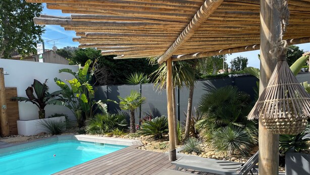 Um deck de madeira com piscina, cercado por plantas tropicais e uma pérgula.