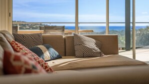 A modern living room with a sofa, patterned cushions, and a large window offering a view of the sea.