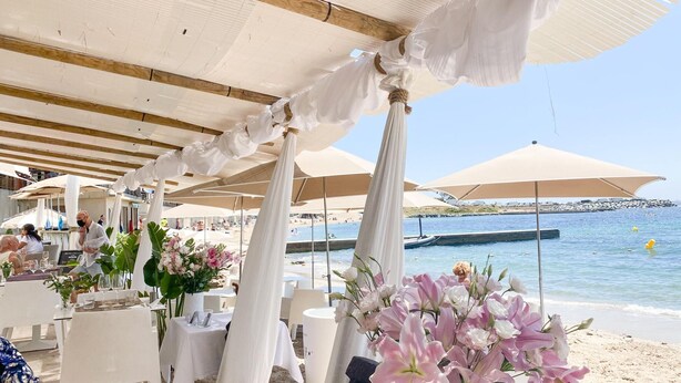 An outdoor seating area with white tables and chairs, arranged under white canopies, overlooking a beach and the sea.