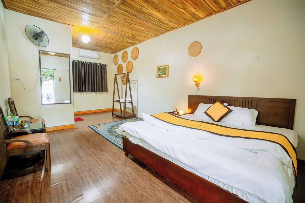 Une chambre d’hôtel avec un plafond en bois, un lit avec une literie blanche et un détail jaune, un bureau avec une chaise, un ventilateur sur pied et un miroir.