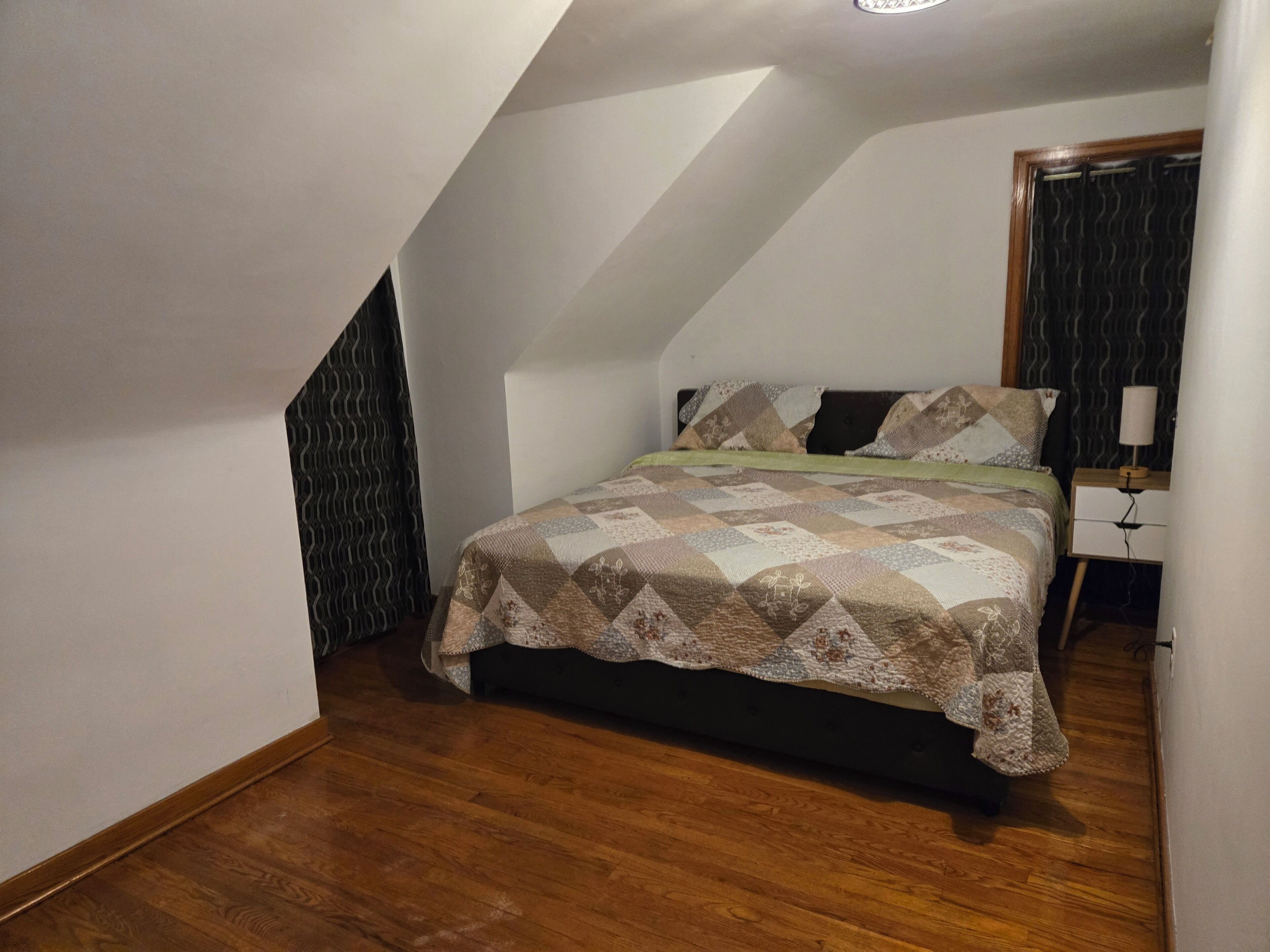 Een slaapkamer met een schuine kap, een bed met een geborduurde quilt, een nachtkastje met een lamp en een houten vloer.