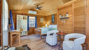 Eine Holzhütte mit einem Bett, zwei Stühlen, einem Tisch, einem Deckenventilator und einem Regal mit Flaschen und Gläsern.