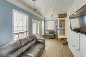 A room with a leather sofa, a small couch, a TV mounted on a brick wall, and a door leading to another room. - Cozy Home Near Table Rock & Eureka Springs (Holiday Island)