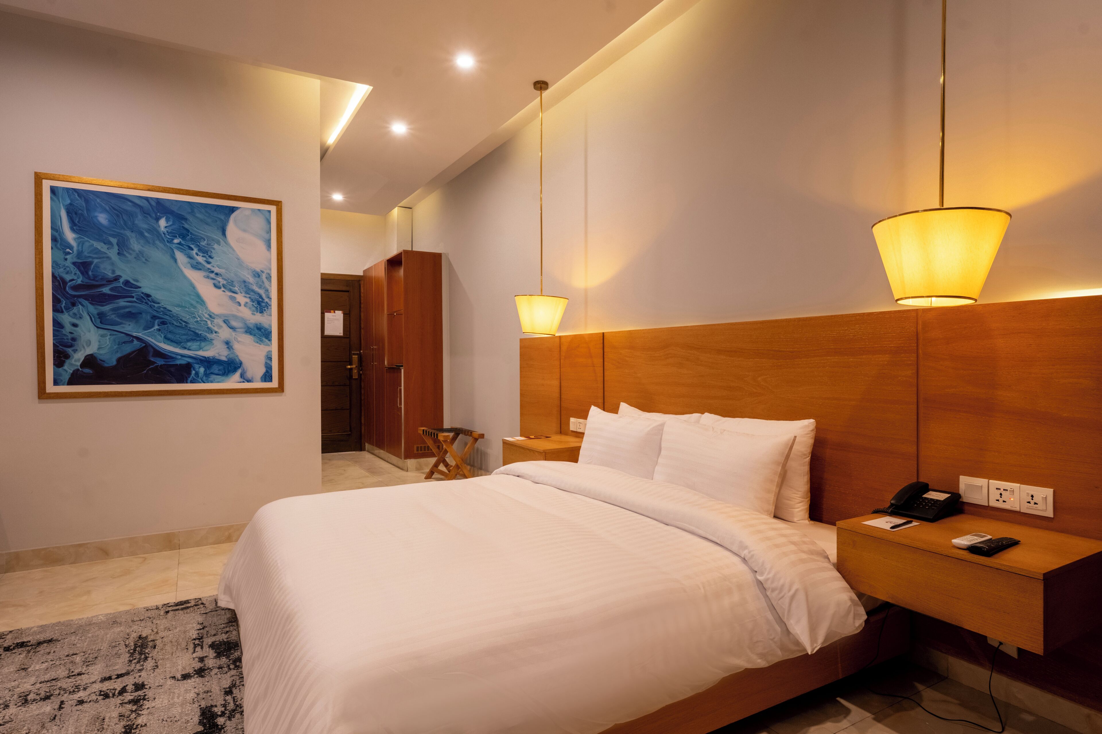 Quarto de hotel com uma cama grande, luminárias de cabeceira, um painel de madeira na cabeceira, mesa de cabeceira com telefone e controle remoto, e um quadro na parede.