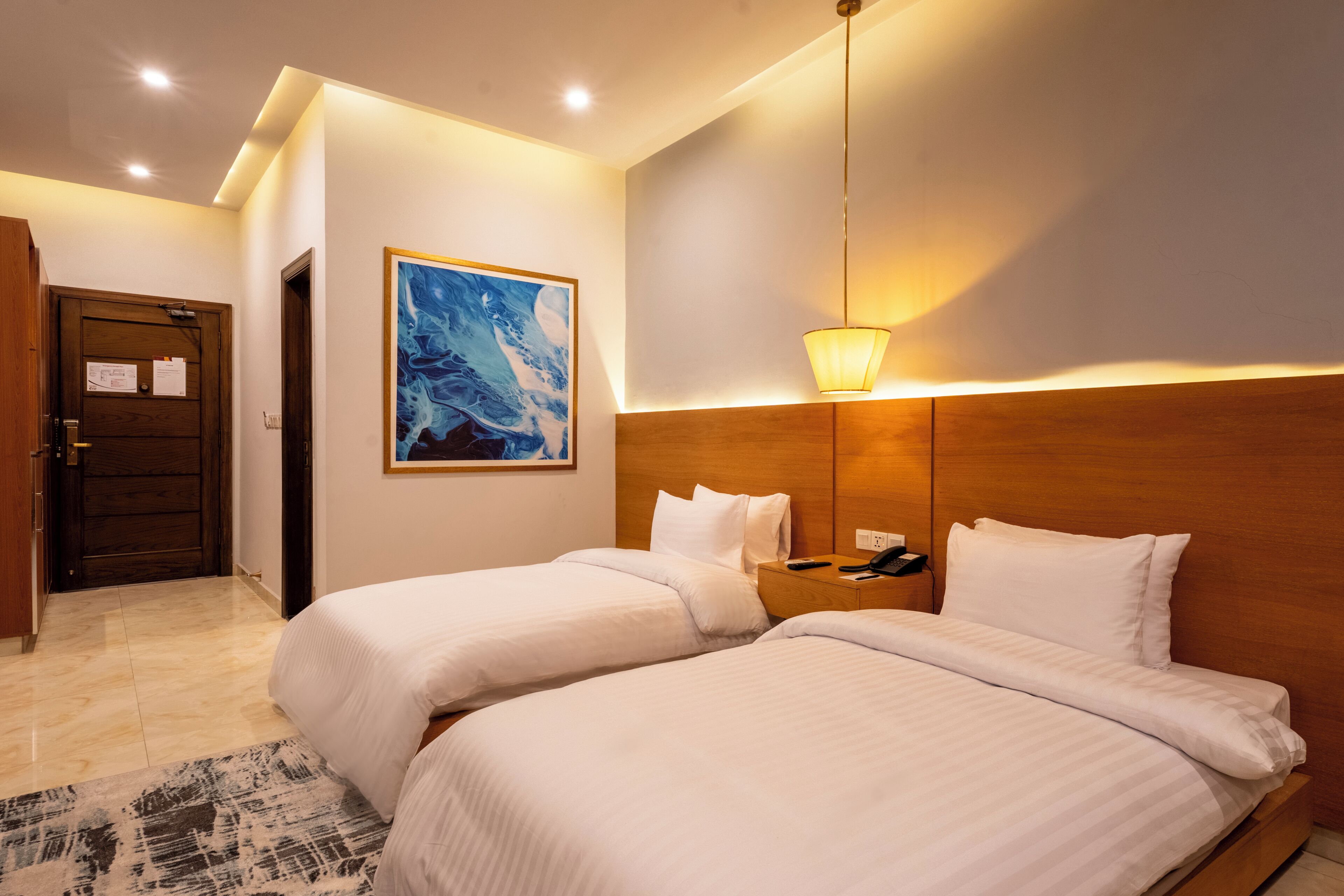 Een hotelkamer met twee bedden, een houten hoofdbord, een schilderij aan de muur, een nachtkastje met een telefoon en een deur met een bericht.
