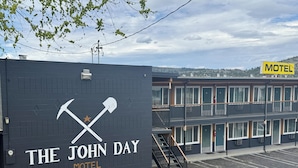 A motel with a distinctive logo featuring crossed tools and a star, a parking lot, and a multi-story building with balconies. - The John Day Motel (John Day)