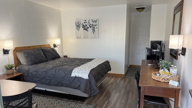 A compact hotel room with a bed, bedside table, chair, desk, and a small kitchenette. - The John Day Motel (John Day)