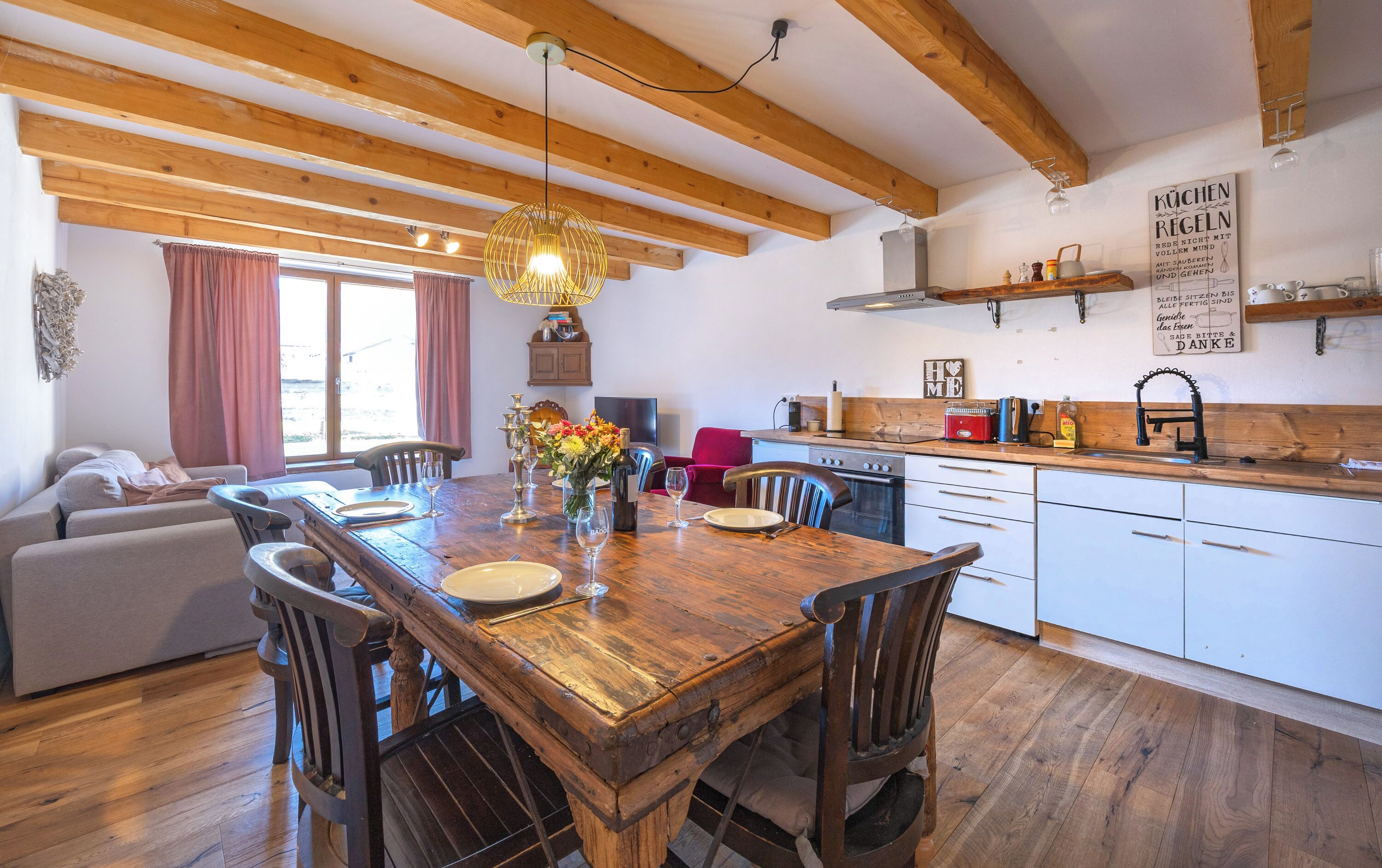 A wooden dining table with chairs, a kitchen with white cabinets, a hanging light fixture, and a window with curtains.