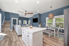 A modern kitchen with a central island, a dining area with a table and chairs, a ceiling fan, recessed lighting, and a view of the outdoors through a window.