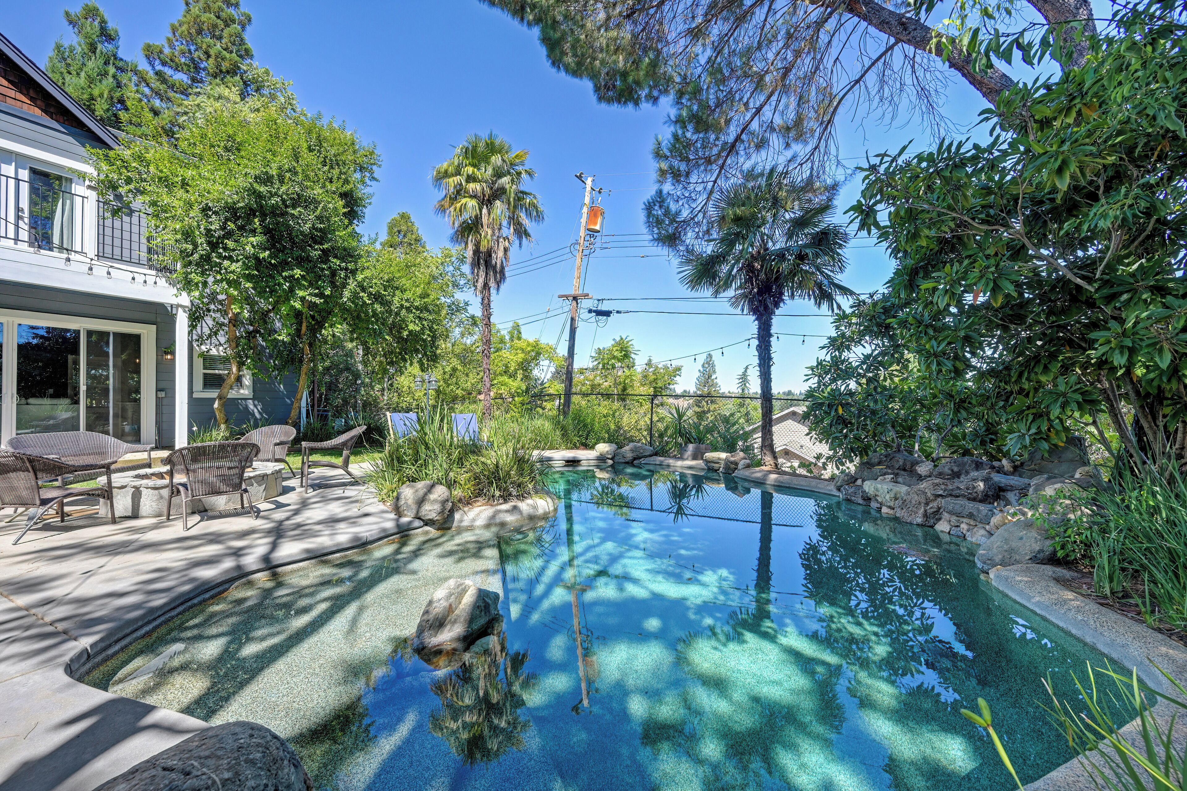 A swimming pool with clear water, surrounded by rocks and plants, and a patio area with chairs and a table.