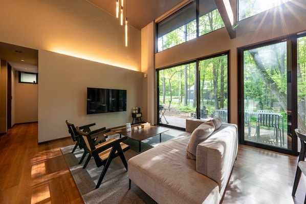 A modern living room with a sofa, coffee table, and chairs, large windows with a forest view, and a flat-screen TV mounted on the wall.