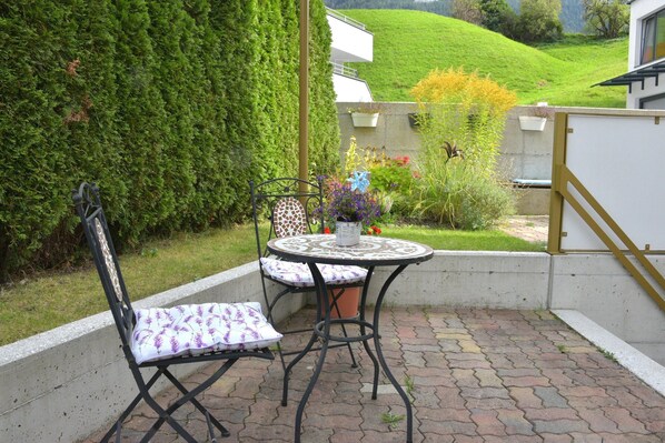 A patio with a table and chairs, surrounded by a hedge and a view of a hill.