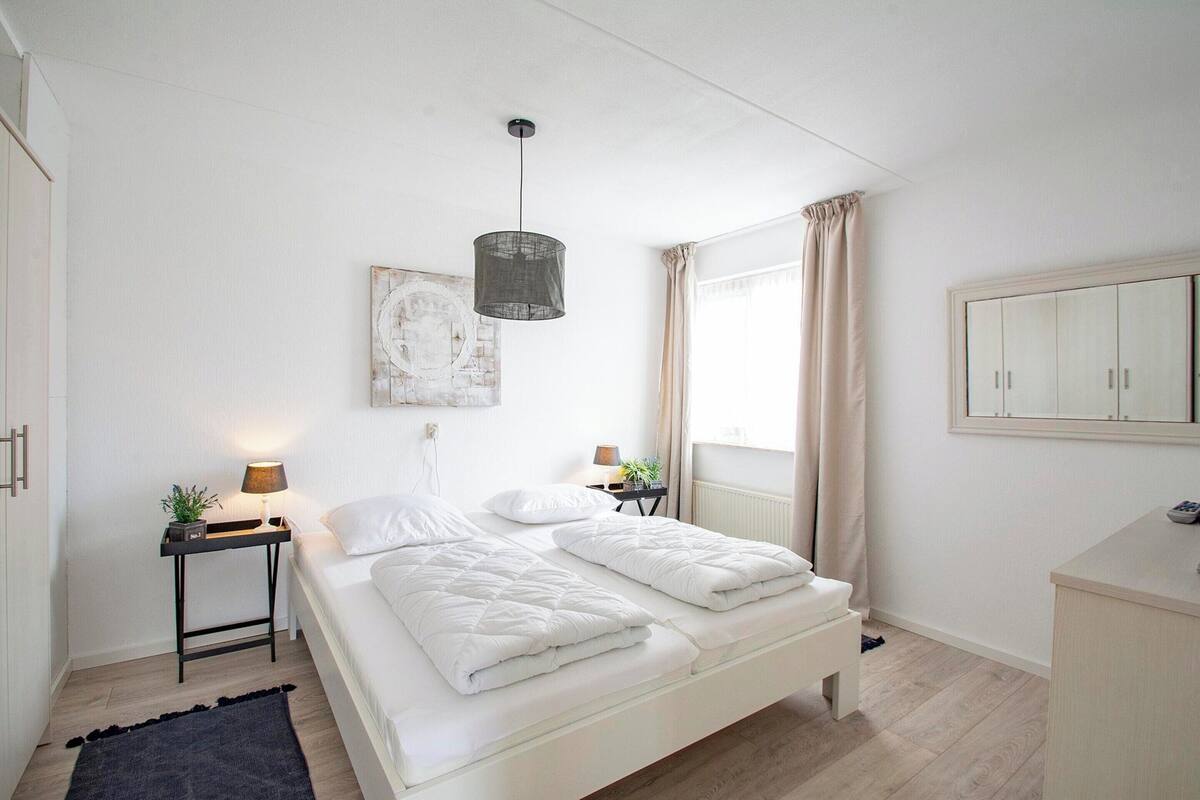 A bedroom with a bed, bedside tables, a hanging light fixture, a window with curtains, and a wardrobe.