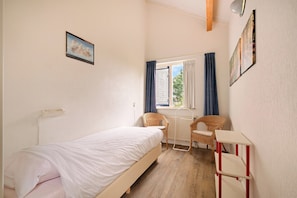 Ein Schlafzimmer mit einem Bett, zwei KorbstĂŒhlen, einem Heizkörper, einem Fenster mit VorhĂ€ngen und einem Wandregal.